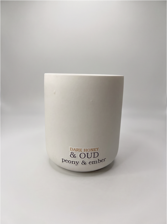 White ceramic candle labelled 'Dark Honey & Oud', a handmade natural vegan wax candle, by Peony & Ember on a warm neutral background