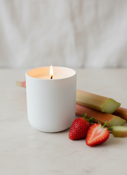 A Peony and Ember natural wax candle in a matte white ceramic vessel placed on a light surface beside fresh rhubarb stalks and ripe strawberries, with soft neutral lighting.